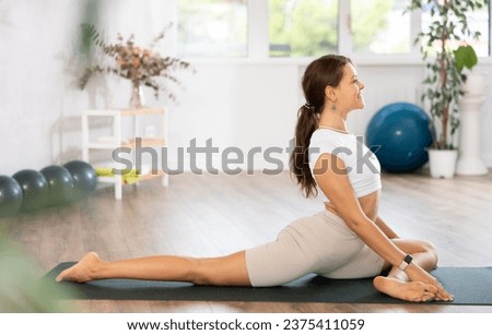 In empty gym hall, girl hatha yoga coach tries to perform various variations of pigeon asana, eka pada rajakapotasana Royalty-Free Stock Photo #2375411059