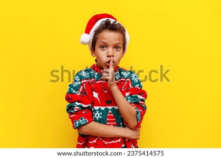 African-American boy in New Year's clothes holds his finger near his lips and shows silence on yellow isolated background, 9-year-old child in Santa hat does not speak