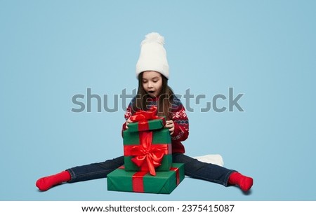 Full body of cute little girl in knitted sweater and hat sitting on stack of gift boxes and opening christmas presents against blue background