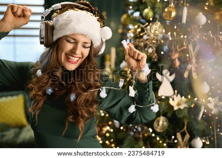 Christmas time. smiling modern 40 years old housewife in green dress listening to the music with headphones and dancing near Christmas tree in the modern house. Royalty-Free Stock Photo #2375484719