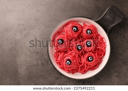 Red pasta with decorative eyes and olives in bowl on grey textured table, top view with space for text. Halloween food