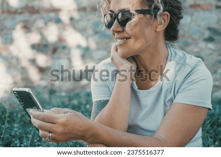 One modern happy woman smile and use mobile phone in outdoor sitting in the garden with green plants in background. Serene female people chatting and sending messages online in outdoor leisure relax Royalty-Free Stock Photo #2375516377