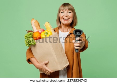 Elderly smiling happy cool woman wearing brown shirt casual clothes hold shopping paper bag with scanning food products check bar code isolated on plain green background. Delivery service from shop