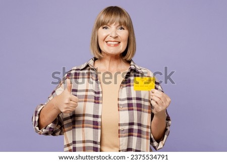 Elderly blonde woman 50s years old wear beige t-shirt shirt casual clothes hold in hand mock up of credit bank card show thumb up isolated on plain pastel light purple background. Lifestyle concept