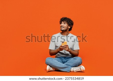 Full body young smiling happy Indian man he wears t-shirt casual clothes sit hold in hand use mobile cell phone look aside isolated on orange red color background studio portrait. Lifestyle concept