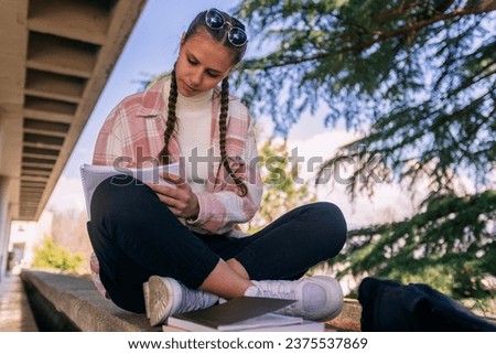 Low angle view photo of beautiful brunette school girl writing homework while sitting on a ledge
