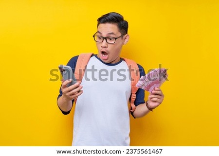 Surprised young Asian student man in casual clothes and glasses backpack holding  mobile phone and cash money isolated on yellow background. Education in High School University College concept Royalty-Free Stock Photo #2375561467
