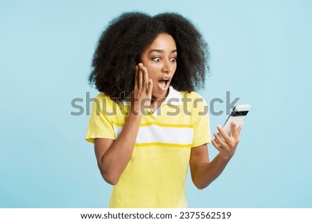 Portrait of excited attractive African American woman wearing casual clothes holding mobile phone, looking at camera isolated on blue background. Concept of online shopping, technology
