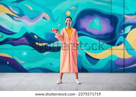 Full length portrait of graffiti woman painter in respirator mask standing near the wall with her paintings looking to camera. Street art concept