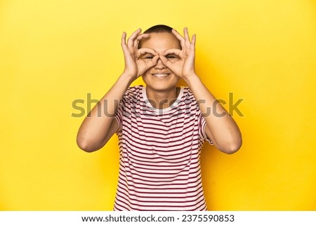 Shaved head woman in red striped tee, yellow backdrop showing okay sign over eyes