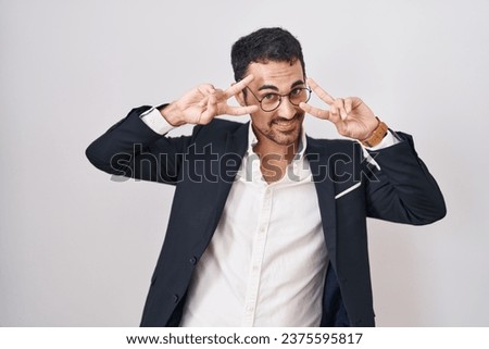 Handsome business hispanic man standing over white background doing peace symbol with fingers over face, smiling cheerful showing victory 