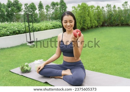 Fitness woman holding yoga mat with Fresh Apple Heathy clean vegan food before working out Beautiful confident asian woman practices in green park . Royalty-Free Stock Photo #2375605373