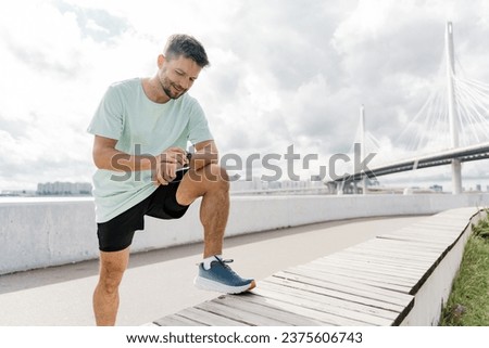 Uses the fitness tracker watch app. A sporty man in a T-shirt training in the open air. Happy guy hobby running instructor before exercises. Royalty-Free Stock Photo #2375606743