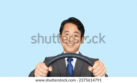 Asian middle-aged man in business suit holding the steering wheel and driving in front of blue background. Royalty-Free Stock Photo #2375614791