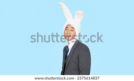 A middle-aged Asian man in a business suit wearing a rabbit headgear in front of a blue background. Party entertainment. Halloween party. Royalty-Free Stock Photo #2375614837