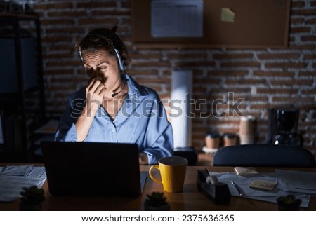 Beautiful brunette woman working at the office at night smelling something stinky and disgusting, intolerable smell, holding breath with fingers on nose. bad smell  Royalty-Free Stock Photo #2375636365