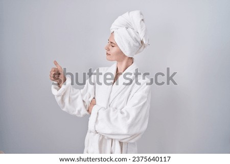 Blonde caucasian woman wearing bathrobe looking proud, smiling doing thumbs up gesture to the side 