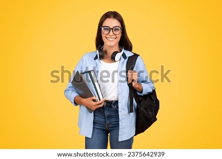 Positive teen student lady with backpack, books, in glasses and headphones study, enjoy learn, isolating on yellow studio background. Education at school, achievement, lifestyle and project