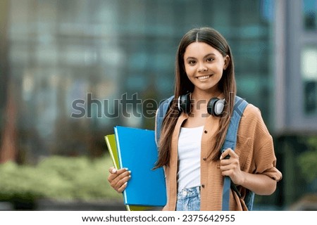 Cheerful student clutching books at sunny campus. Happy young woman gripping books at vibrant campus, exuding positivity and youthful academic spirit, carrying backpack, copy space Royalty-Free Stock Photo #2375642955
