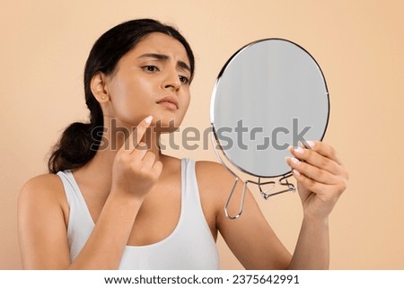 Skin Problems Concept. Unhappy Young Indian Woman Holding Magnifying Mirror And Looking At Pimple On Her Chin, Upset Eastern Female Inspecting Acne, Standing Isolated Over Beige Studio Background Royalty-Free Stock Photo #2375642991