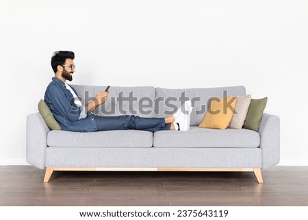 A young Indian man enjoys leisure time, reclining comfortably on a grey couch, engaging with his smartphone, isolated on a white background, portraying relaxation and digital lifestyle, copy space Royalty-Free Stock Photo #2375643119