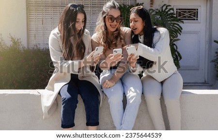 Three young women sitting on the street looking at their smartphones, opening their mouths in surprise. Youth concept. Royalty-Free Stock Photo #2375644539