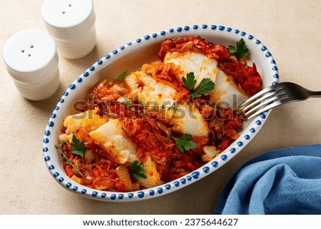Served Greek and Polish style oven baked cod fish Psari Plaki with sauteed, shredded vegetables, carrot, onion, parsley and tomato sauce. White plate with blue decoration. Beige background.