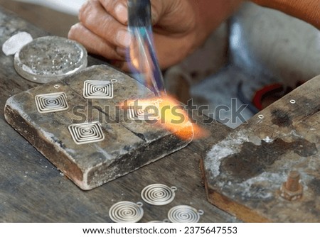 The goldsmith designs rounded and squared alloy silver pendants, a creative craftsman manufacturing jewelry, close-up on his hands holding pliers and the burner with a flame. Royalty-Free Stock Photo #2375647553