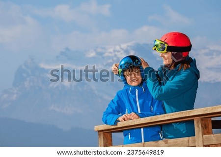 Mother putting ski helmet on the head of her son on wooden hotel balcony, ready to ride