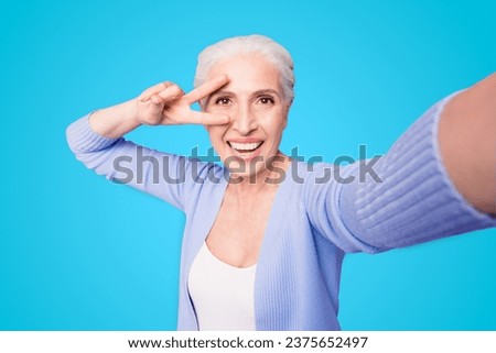 Elderly lady makes selfie on the camera of a smartphone making v-sign near the eyes isolated on bright violet background with copy space for text