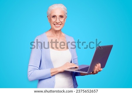 Grey haired old nice beautiful cheerful smiling woman wearing spectacles holding laptop learning online course. Isolated over violet purple background