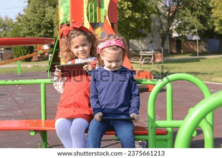 Two little girls use a tablet on the playground. Small children playing outdoors