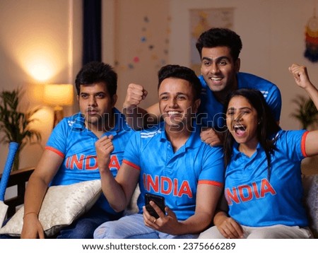 Thrilled friends in Indian jerseys cheering for team India during match. Excited cricket fans sitting at home watching game on TV, using smartphone app to online bet, friends celebrating victory