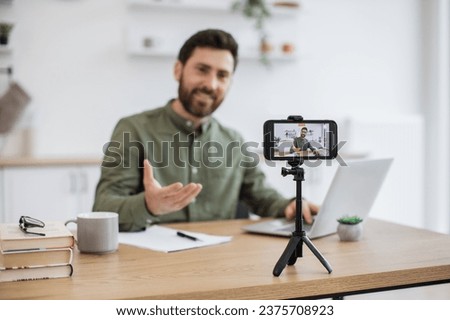 Modern smartphone fixed on tripod filming handsome caucasian man in casual wear working on portable laptop at home office. Blurred background. Technology, people and blogging concept.