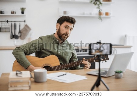 Inspired caucasian man virtuoso playing guitar while sitting at desk and recording video on modern smartphone with tripod. Male vlogger creating new lesson about instrument techniques to social media. Royalty-Free Stock Photo #2375709013