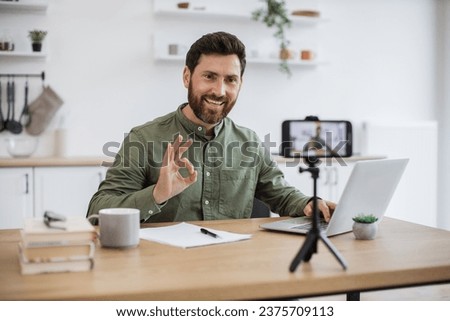 Confident bearded man sitting at desk and working on wireless laptop while recording video on modern smartphone fixed on tripod showing sign ok