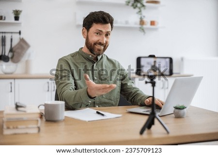 Confident bearded man sitting at desk and working on wireless laptop while recording video on modern smartphone fixed on tripod. Male freelancer creating new content for social media at home.