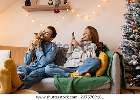 Loved ones. Happy young couple and their small dog together at home during the Christmas holidays. Woman takes a photo of her boyfriend kissing their dog.