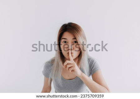 Asian Thai woman wear grey, hold fingers on lips, showing shh, hush quiet sign, making gesture silence, keep secret, looking at camera, isolated on white background wall.