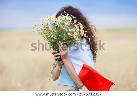Woman with flag of Poland on shoulders holding bouquet of daisies in front of face in wheat field. Polish Flag Day. Independence Day. Travel and learn polish language concept. Selective focus. Royalty-Free Stock Photo #2375712337