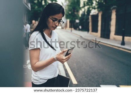 Serious young woman using smartphone while standing at sidewalk in city