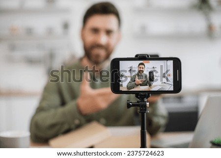 Focus on wireless smartphone standing on tripod and recording video. Cheerful male blogger showing and telling about modern gadget to subscribers in living room interior.