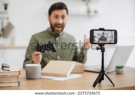 Overjoyed caucasian influencer with modern joystick in hands unboxing new parcel while livestreaming. Online-working middle-aged man filming by fixed modern smartphone on blurred background.
