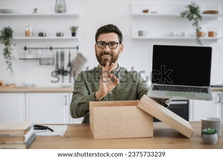 Mobile web camera view of young man in casual attire doing unpacking of new portable laptop while working from home showing sign ok