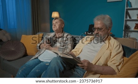 In the picture the elderly couple sit on the sofa in the apartment against the blue wall. The man reads the book aloud. The woman listens, rejoices and drinks tea. Home coziness.