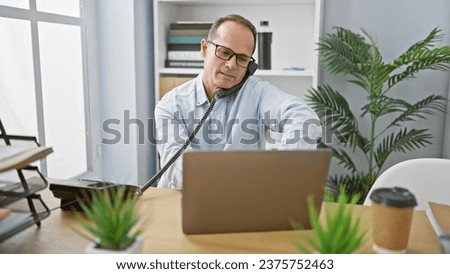 Relaxed middle age businessman engaging in a serious phone conversation while working on his laptop at the office