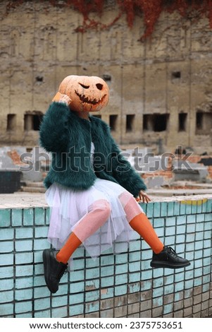 Halloween. a girl in a violet-blue dress, a green fur coat and orange tights and with a pumpkin instead of a head stands and sits on a blue brick wall. Halloween costume. ruins in the background