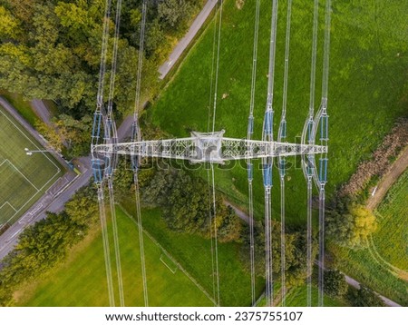 High voltage pylon with power lines between nature and sports field photographed directly from above with drone