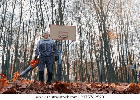 Handsome senior man removing dry leaves with leaf blower on basketball court outside. Low angle view of smiling bearded male worker with portable leaf blower working in morning. Seasonal work concept.