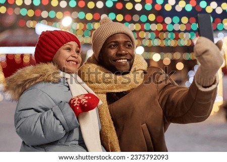 Black man and daughter taking selfie photo outdoors together in Christmas market and smiling, copy space